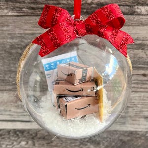 May include: Clear glass Christmas ornament filled with miniature Amazon boxes and white filler. The ornament is topped with a red glittery bow and a red ribbon for hanging. The ornament is a unique holiday decoration.