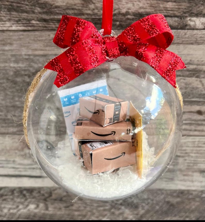 May include: Clear glass Christmas ornament filled with miniature Amazon boxes and white filler. The ornament is topped with a red glittery bow and a red ribbon for hanging. The ornament is a unique holiday decoration.