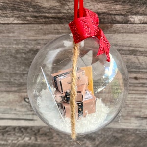 May include: Clear glass Christmas ornament with miniature Amazon boxes inside, resting on white faux snow. The ornament has a red glittery bow and a jute string for hanging. The background is a wooden surface.