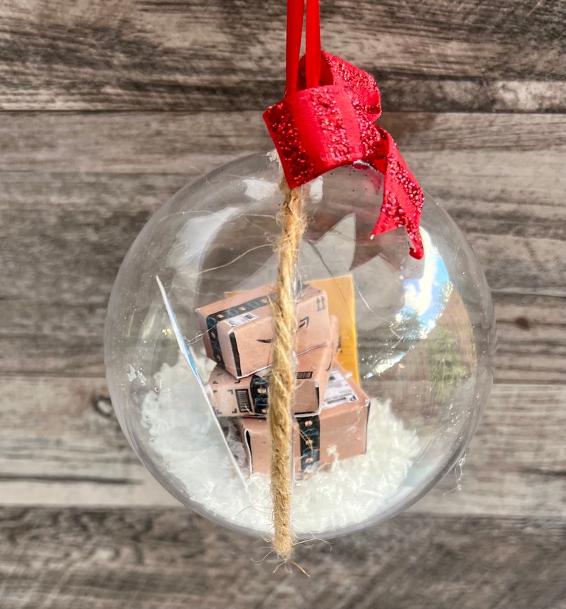 May include: Clear glass Christmas ornament with miniature Amazon boxes inside, resting on white faux snow. The ornament has a red glittery bow and a jute string for hanging. The background is a wooden surface.