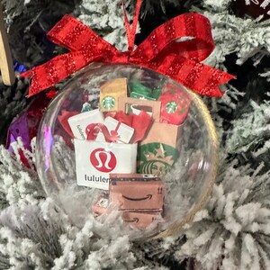May include: A clear glass Christmas ornament filled with miniature shopping bags and gift boxes. The ornament includes a red bow and features logos from Lululemon, Starbucks, and Amazon. The ornament hangs on a snow-covered Christmas tree.