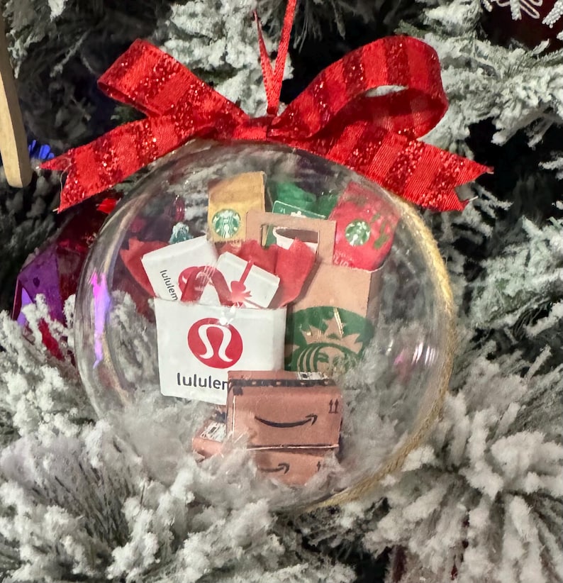 May include: A clear glass Christmas ornament filled with miniature shopping bags and gift boxes. The ornament includes a red bow and features logos from Lululemon, Starbucks, and Amazon. The ornament hangs on a snow-covered Christmas tree.