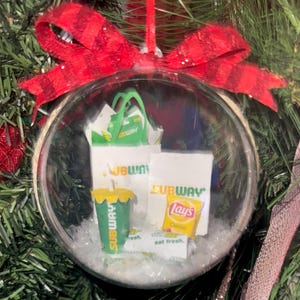 May include: A clear, spherical Christmas ornament containing miniature Subway-branded items, including a white paper bag with green handles, a green drink cup, a yellow Lay's potato chip bag, and a white wrapper, all topped with a red plaid ribbon.