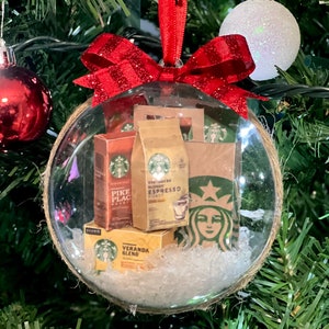 May include: A clear glass Christmas ornament with a red and white striped ribbon bow. Inside the ornament are miniature boxes of Starbucks coffee, including Pike Place Roast, Blonde Espresso Roast, and Veranda Blend.