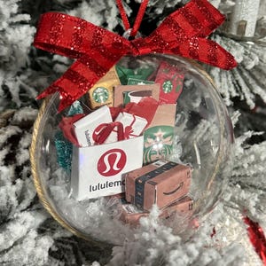 May include: A clear glass Christmas ornament filled with miniature shopping bags from Lululemon, Starbucks, and Amazon. The ornament is topped with a red glittery bow and hangs from a red string. The background is a snowy Christmas tree.