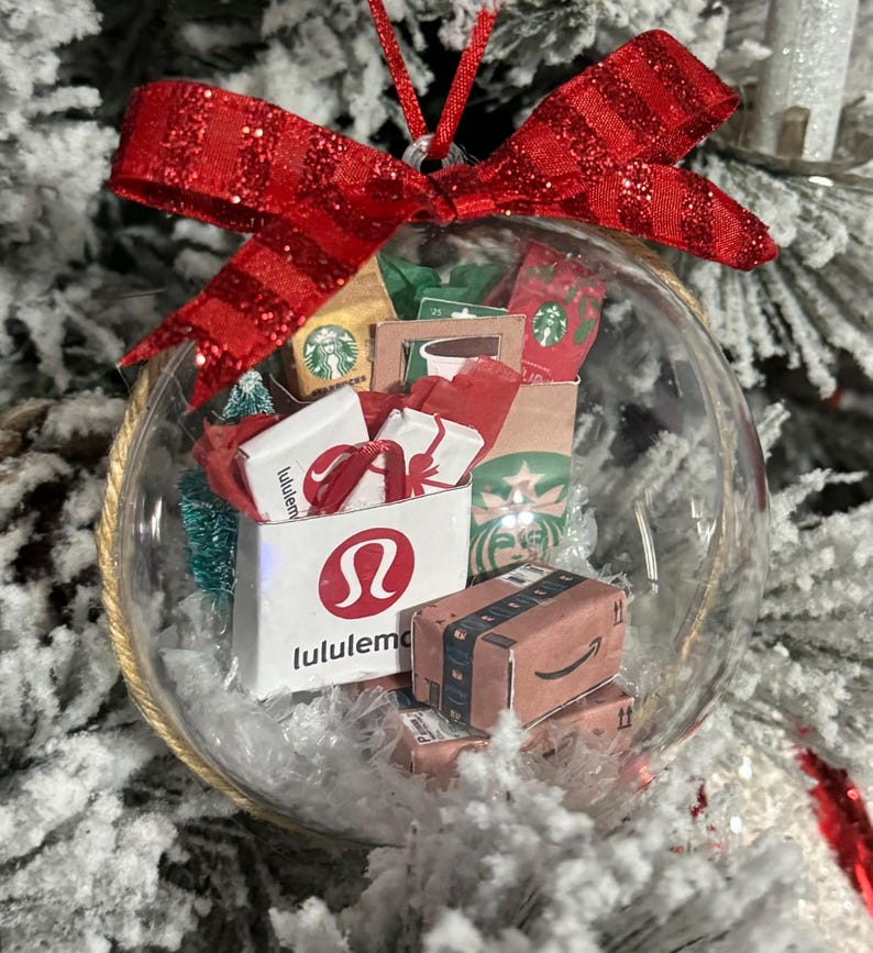 May include: A clear glass Christmas ornament filled with miniature shopping bags from Lululemon, Starbucks, and Amazon. The ornament is topped with a red glittery bow and hangs from a red string. The background is a snowy Christmas tree.
