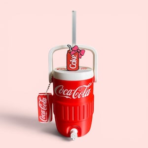 May include: A red and white Coca-Cola beverage cooler with a white handle, a spigot at the base, and a clear straw. A small Coca-Cola can charm hangs from a chain on the handle, and a decorative red can topper with a pink bow sits on the lid.