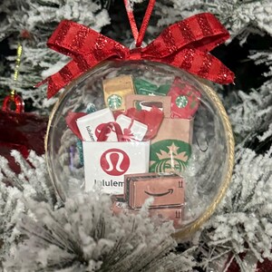 May include: A clear Christmas ornament filled with miniature shopping bags and boxes from Lululemon, Starbucks, and Amazon. The ornament is topped with a red bow and hangs from a red ribbon, set against a snowy Christmas tree.