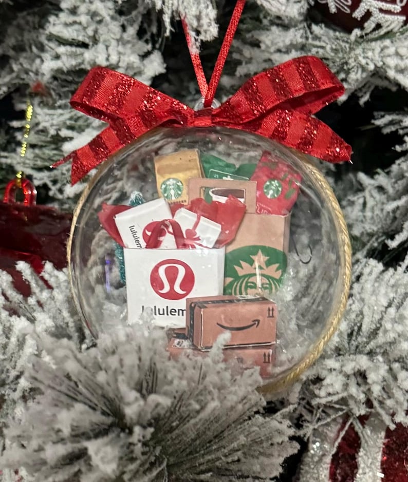 May include: A clear Christmas ornament filled with miniature shopping bags and boxes from Lululemon, Starbucks, and Amazon. The ornament is topped with a red bow and hangs from a red ribbon, set against a snowy Christmas tree.