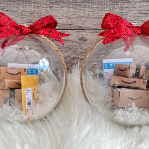 May include: Two clear Christmas ornaments filled with miniature Amazon boxes and packing materials. Each ornament is topped with a red ribbon bow and has a jute rope loop for hanging. The ornaments sit on a white faux fur surface.