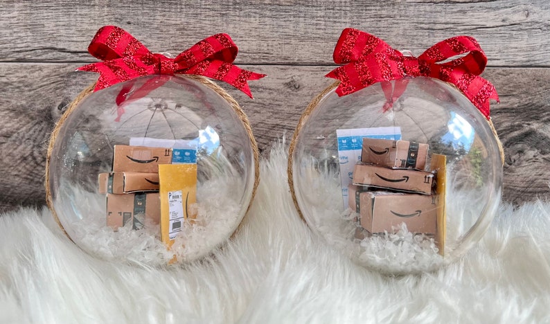 May include: Two clear Christmas ornaments filled with miniature Amazon boxes and packing materials. Each ornament is topped with a red ribbon bow and has a jute rope loop for hanging. The ornaments sit on a white faux fur surface.