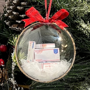 May include: A clear glass Christmas ornament with a red bow and a stack of miniature Priority Mail boxes inside. The ornament is surrounded by white shredded paper.