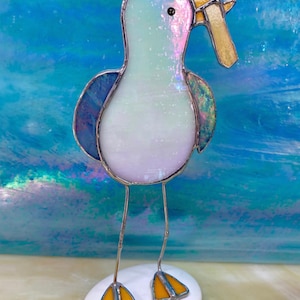 Könnte beinhalten: Eine Glasmalerei-Möwen-Skulptur mit hellblauem und schillerndem Körper, blauen Flügeln und gelben Füßen. Die Möwe hält ein gelbes und goldenes Objekt in ihrem Schnabel. Die Skulptur steht auf einem weißen Stein vor blauem Hintergrund.