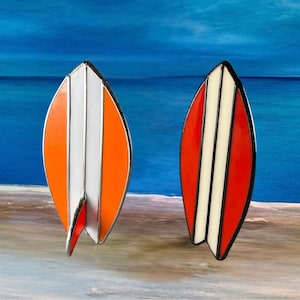 Puede incluir: Dos broches con forma de tabla de surf. Uno es naranja con rayas blancas, el otro es rojo con rayas crema. Ambos tienen un contorno plateado y una pequeña aleta. El fondo es una escena de playa con agua y cielo azules.