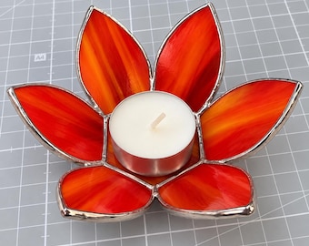 Könnte beinhalten: Ein Kerzenhalter aus Buntglas in Form einer Blume mit sechs Blütenblättern in Orange- und Rottönen. Die Blütenblätter sind durch silberfarbene Metalllötstellen verbunden. Ein weißes, rundes Teelicht steht in der Mitte der blütenförmigen Basis.