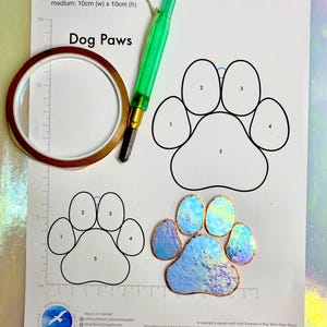May include: A printable pattern for a dog paw shaped stained glass project. The pattern includes five sections numbered 1 through 5. The pattern is printed on white paper with a ruler and measurements in centimeters. A finished stained glass paw is shown in the bottom right corner of the image.
