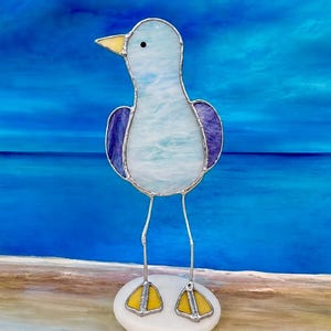 Pu&ograve; includere: Scultura di gabbiano in vetro colorato con corpo azzurro, ali viola e becco e zampe gialle. L'uccello &egrave; in piedi su una base ovale bianca con gambe di filo d'argento. Lo sfondo &egrave; una scena oceanica blu sfocata.
