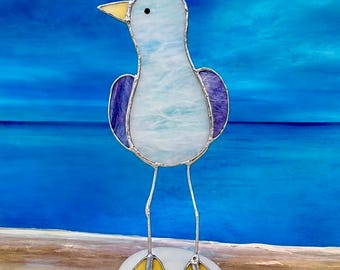 Könnte beinhalten: Eine Glasmalerei-Möwen-Skulptur mit hellblauem Körper, lila Flügeln und gelbem Schnabel und Füßen. Der Vogel steht auf einem weißen, ovalen Sockel mit silbernen Drahtbeinen. Der Hintergrund ist eine verschwommene blaue Meereslandschaft.