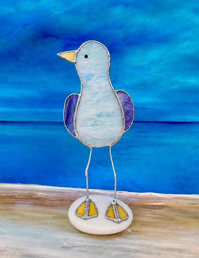 Pu&ograve; includere: Scultura di gabbiano in vetro colorato con corpo azzurro, ali viola e becco e zampe gialle. L'uccello &egrave; in piedi su una base ovale bianca con gambe di filo d'argento. Lo sfondo &egrave; una scena oceanica blu sfocata.
