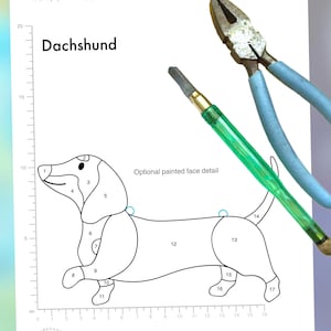 Peut inclure: Un patron de chien teckel, mesurant 17 cm (l) x 11 cm (h), avec un schéma numéroté. L'image présente également des outils, tels que des pinces et un coupe-verre, pour le travail du verre.