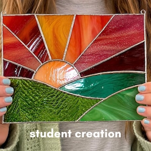 Op de afbeelding: Gebrandschilderd glaspaneel met een zonsondergang boven een landschap. De zon is een halve cirkel van oranje glas, met rode, oranje en gele stralen. Het landschap heeft groen getextureerd glas. De tekst "student creation" staat onderaan.