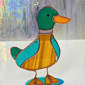 May include: A stained glass duck figurine with a teal body, yellow wings, and a green head. The duck is standing on a teal base.