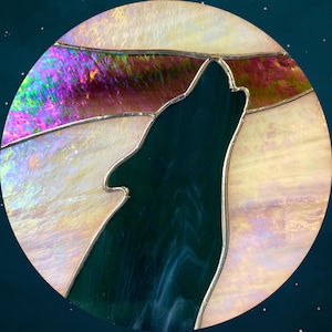 May include: Stained glass artwork depicting a howling wolf silhouette against a full moon. The moon features iridescent glass with shades of purple, pink, and yellow. The wolf is a dark teal color, and the piece is framed with a silver solder.