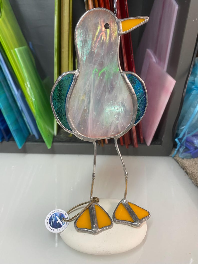 Pu&ograve; includere: Una scultura di gabbiano in vetro colorato con corpo perlato, ali turchesi e becco e zampe gialle. L'uccello &egrave; in piedi su una base di pietra bianca. La scultura ha sottili gambe di metallo e una piccola etichetta attaccata.