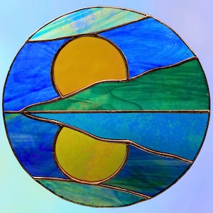 Puede incluir: Obra de arte circular de vidrieras con un sol amarillo y su reflejo. El diseño incorpora vidrio azul, verde y amarillo, enmarcado por un borde color cobre. La obra representa un paisaje con un sol y su reflejo.
