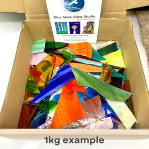 May include: A cardboard box filled with assorted stained glass scraps in various shapes, sizes, and colours including blue, orange, green, yellow, and white. A Blue Skies Glass Studio card is inside the box, and the text 1kg example is printed below.