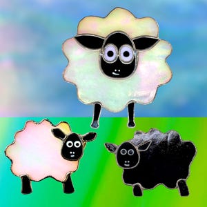 Op de afbeelding: Drie decoratieve schapenbeeldjes met zwarte gezichten en poten. Eén schaap is wit, één is roze en één is zwart. De schapen hebben een parelmoerachtige, getextureerde uitstraling tegen een blauw-groene gradiënt achtergrond.
