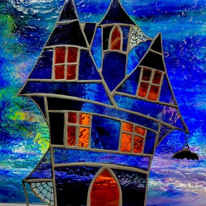 May include: Stained glass haunted house in blue and black. The windows are orange and red, and a bat hangs from the side. A spider web is visible. A Halloween decoration, suitable for display.