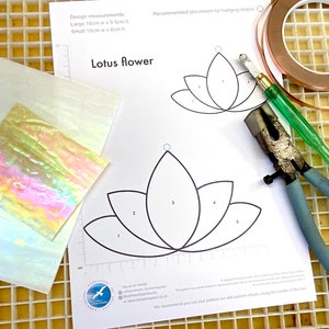 Op de afbeelding: Een wit vel papier met een zwart-witte omtrek van een lotusbloem. Het papier is gelabeld "Lotus Flower" en bevat instructies voor het uitsnijden van de bloem. De afbeelding toont ook een stuk iriserend glas, koperfolietape, een glassnijder en een tang.
