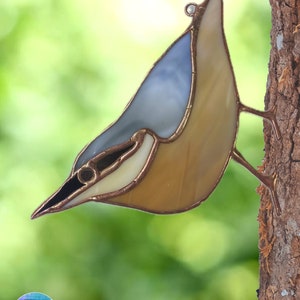 以下が含まれることがあります： 青、茶色、白のガラスでできたステンドグラスの鳥。鳥は木の枝にとまっている。