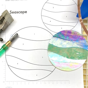 May include: A black and white pattern for a seascape stained glass project. The pattern includes two circles, one large and one small, with wavy lines to represent the ocean. The pattern is labelled with numbers 1 through 5 to indicate the different pieces of glass needed for the project. The text "Seascape" is at the top of the pattern.