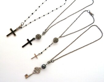 May include: Three silver necklaces with different pendants. The first necklace has a black cross pendant. The second necklace has a black cross pendant and a small silver ball. The third necklace has a silver key pendant, a small silver ball, and a black pearl.