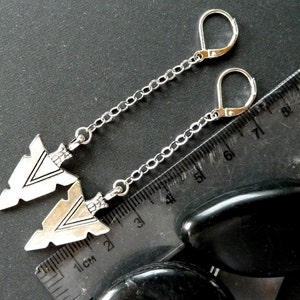 Op de afbeelding: Zilverkleurige oorbellen met pijlpunthangers. De oorbellen hebben een beugel sluiting en hangen aan een fijne ketting. De pijlen hebben ge&euml;tste details en worden getoond op een donkere ondergrond met een liniaal.
