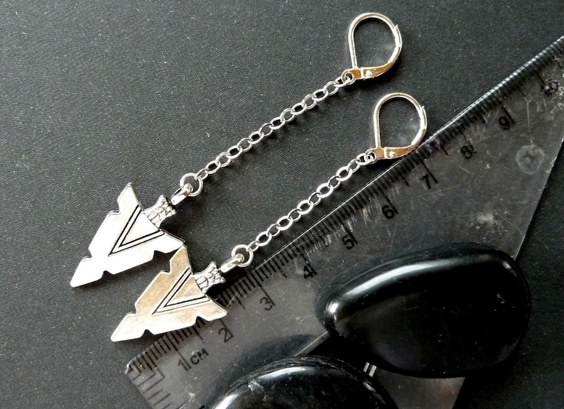 Op de afbeelding: Zilverkleurige oorbellen met pijlpunthangers. De oorbellen hebben een beugel sluiting en hangen aan een fijne ketting. De pijlen hebben ge&euml;tste details en worden getoond op een donkere ondergrond met een liniaal.