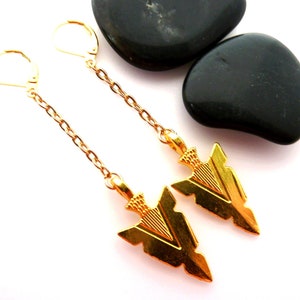 Op de afbeelding: Een paar goudkleurige oorbellen met een pijlontwerp. De oorbellen hebben een ketting en een sluiting met hefboom. De pijlen zijn gedetailleerd met ge&euml;tste lijnen en worden gepresenteerd op een wit oppervlak met twee zwarte stenen.
