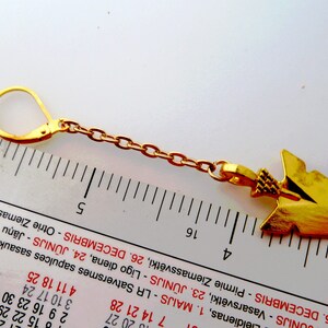 Op de afbeelding: Goudkleurige oorbel met een pijlontwerp. De oorbel heeft een oorhaaksluiting en een ketting. De pijlhanger is ongeveer 4 cm lang. De oorbel wordt getoond naast een liniaal.