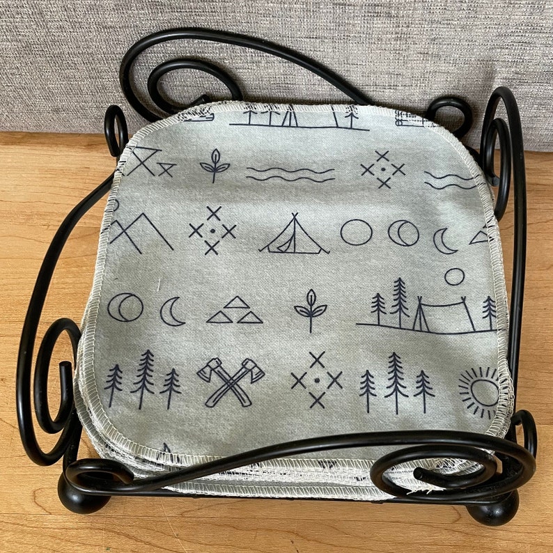 May include: A stack of gray cotton dishcloths with a black and white camping themed pattern. The dishcloths are folded and stacked in a black metal holder.