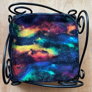 May include: A stack of colorful tie-dye fabric squares with a black background. The fabric has a galaxy-like pattern with shades of yellow, orange, red, blue, and green. The fabric is displayed on a black metal stand.