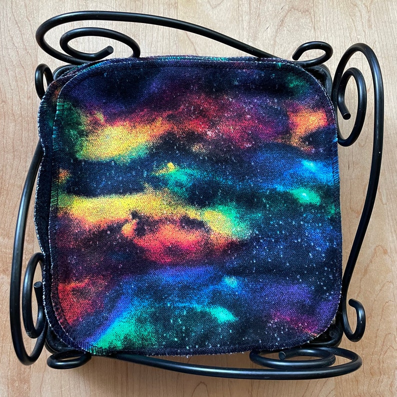 May include: A stack of colorful tie-dye fabric squares with a black background. The fabric has a galaxy-like pattern with shades of yellow, orange, red, blue, and green. The fabric is displayed on a black metal stand.