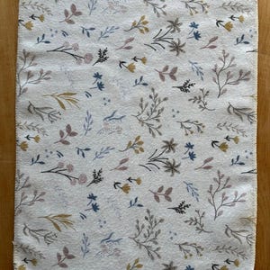 May include: A rectangular off-white fabric cloth with rounded corners featuring a repeating pattern of small botanical illustrations including sprigs, leaves, and flowers in muted shades of blue, yellow, gray, and mauve. The text Wild herbs is visible.