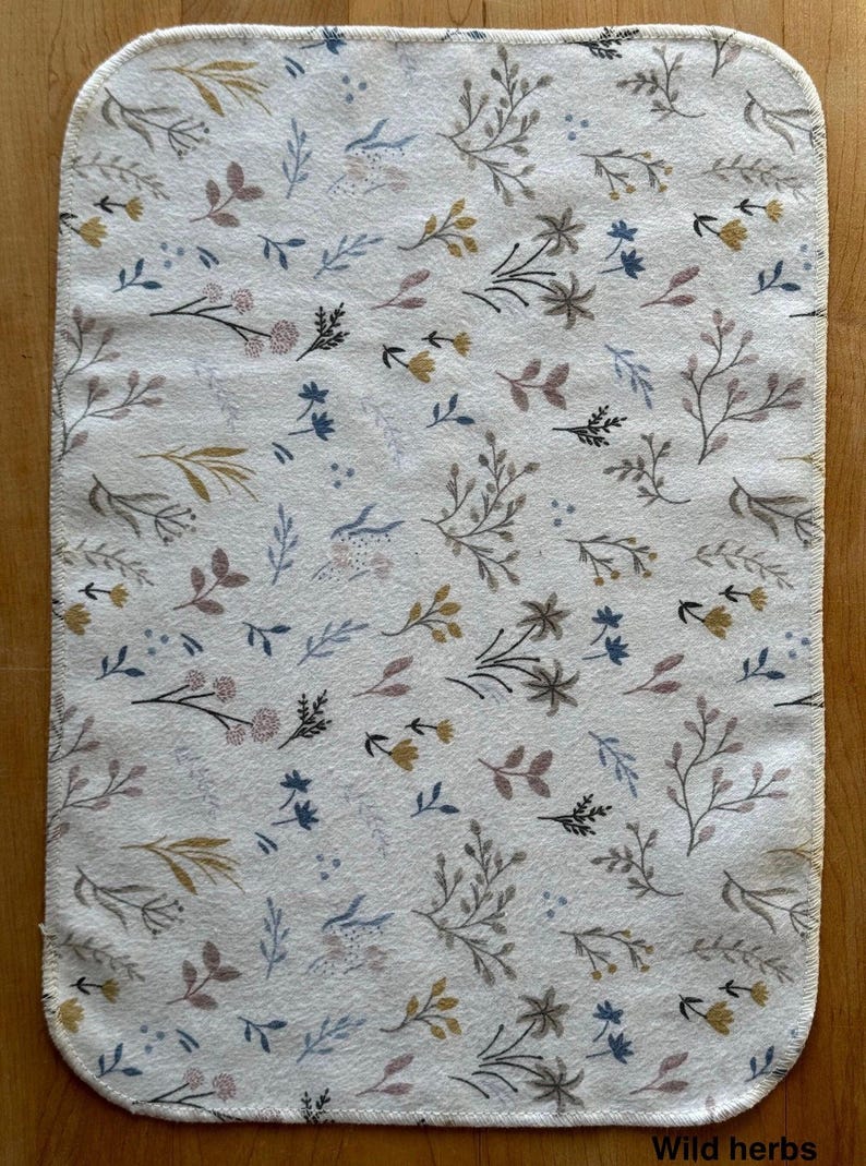 May include: A rectangular off-white fabric cloth with rounded corners featuring a repeating pattern of small botanical illustrations including sprigs, leaves, and flowers in muted shades of blue, yellow, gray, and mauve. The text Wild herbs is visible.
