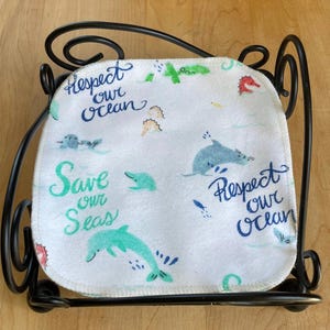 Pode incluir: Um pano de tecido branco quadrado com cantos arredondados apresentando um padr&atilde;o de golfinhos, criaturas marinhas e o texto "Respect our ocean" e "Save our seas" em azul e verde, contido num suporte de arame preto com detalhes em volutas.