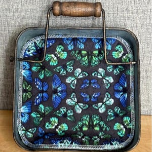 May include: A square metal tray with a dark, weathered finish and a wooden handle, containing a fabric square featuring a repeating pattern of blue and green butterflies on a black background, with white stitched edges and the text Butterflies/ BL.