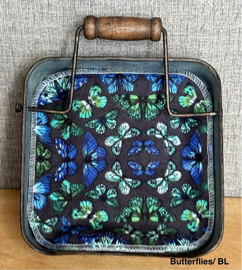 May include: A square metal tray with a dark, weathered finish and a wooden handle, containing a fabric square featuring a repeating pattern of blue and green butterflies on a black background, with white stitched edges and the text Butterflies/ BL.