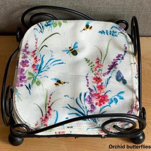May include: A stack of white square fabric cloths featuring a colorful print of bees, butterflies, and floral stems in shades of pink, purple, and blue, held within a black metal wire holder with decorative scrollwork at the corners.