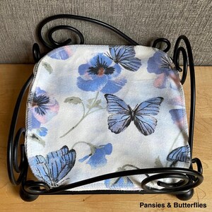 May include: A black metal napkin holder with a square base and scrolled wire corners, containing a square fabric napkin featuring a light blue background with a pattern of blue pansy flowers and blue butterflies, labeled Pansies & Butterflies.